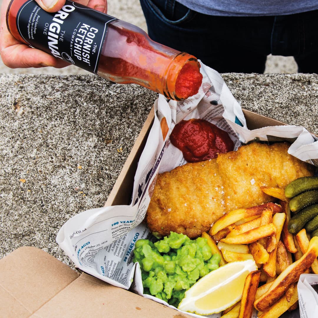 Cornish ketchup poured onto fish, chips and mushy peas in a takeaway box