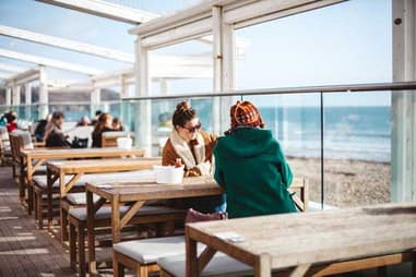 Customers at wooden tables on a glass-fronted beachside café terrace