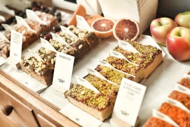 Labelled traybakes on a deli counter beside apples and blood oranges