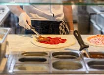Gloved catering worker spreading tomato sauce on pizza bases