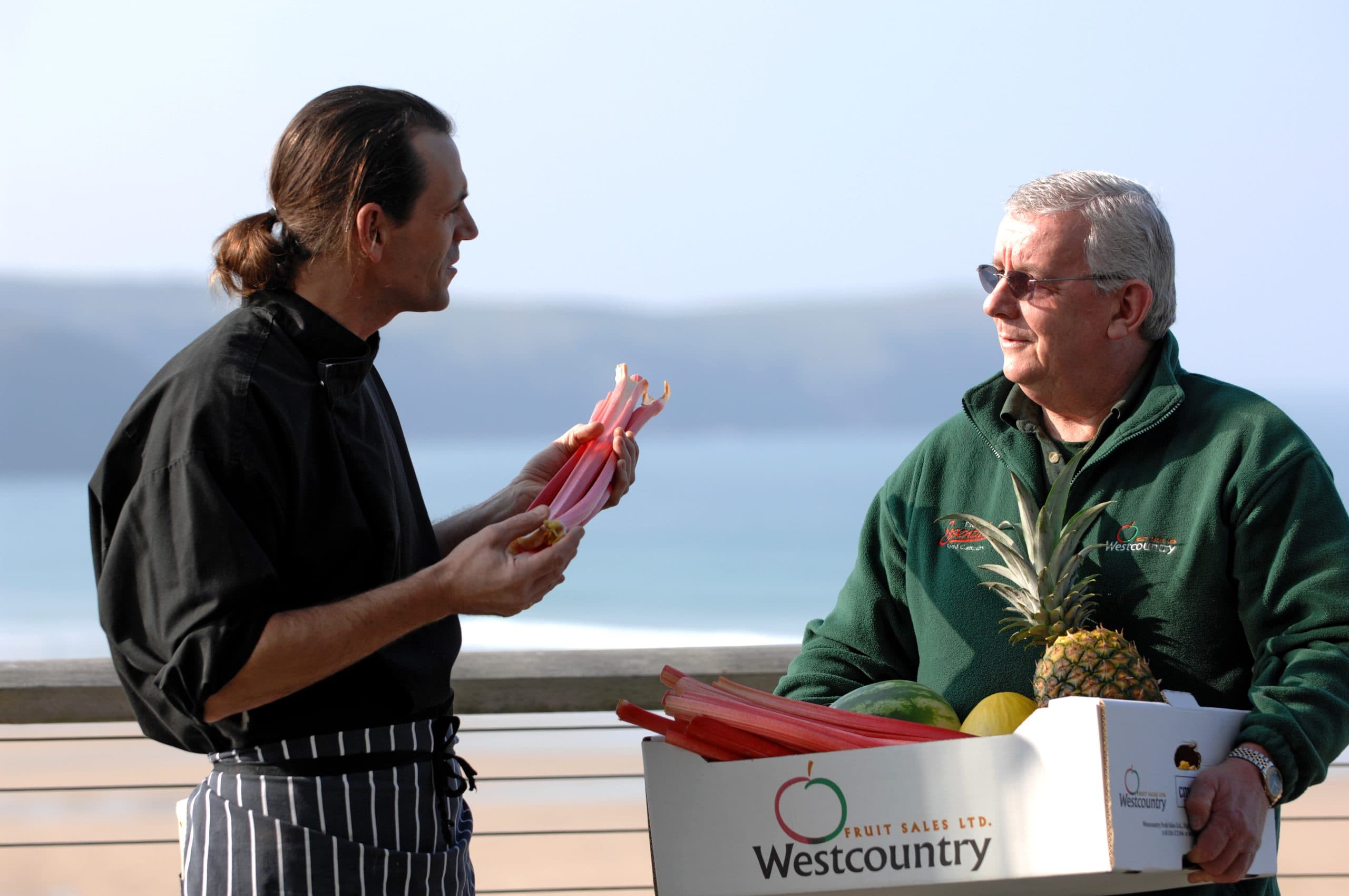 Chef holding rhubarb beside a man with a box of Westcountry fruit by the sea