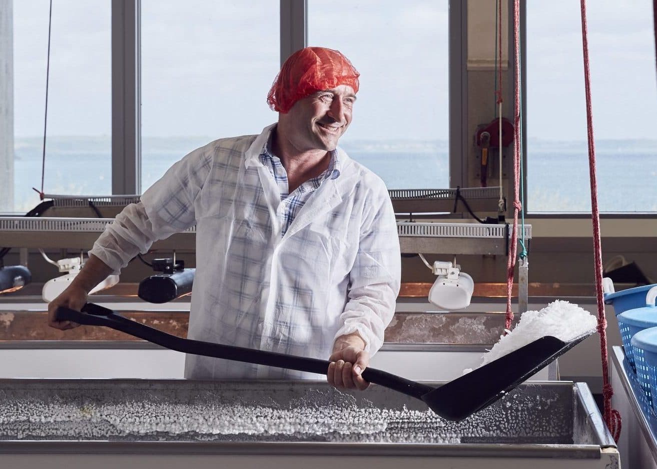 Man in a hairnet hand-harvesting sea salt crystals with a shovel by the sea