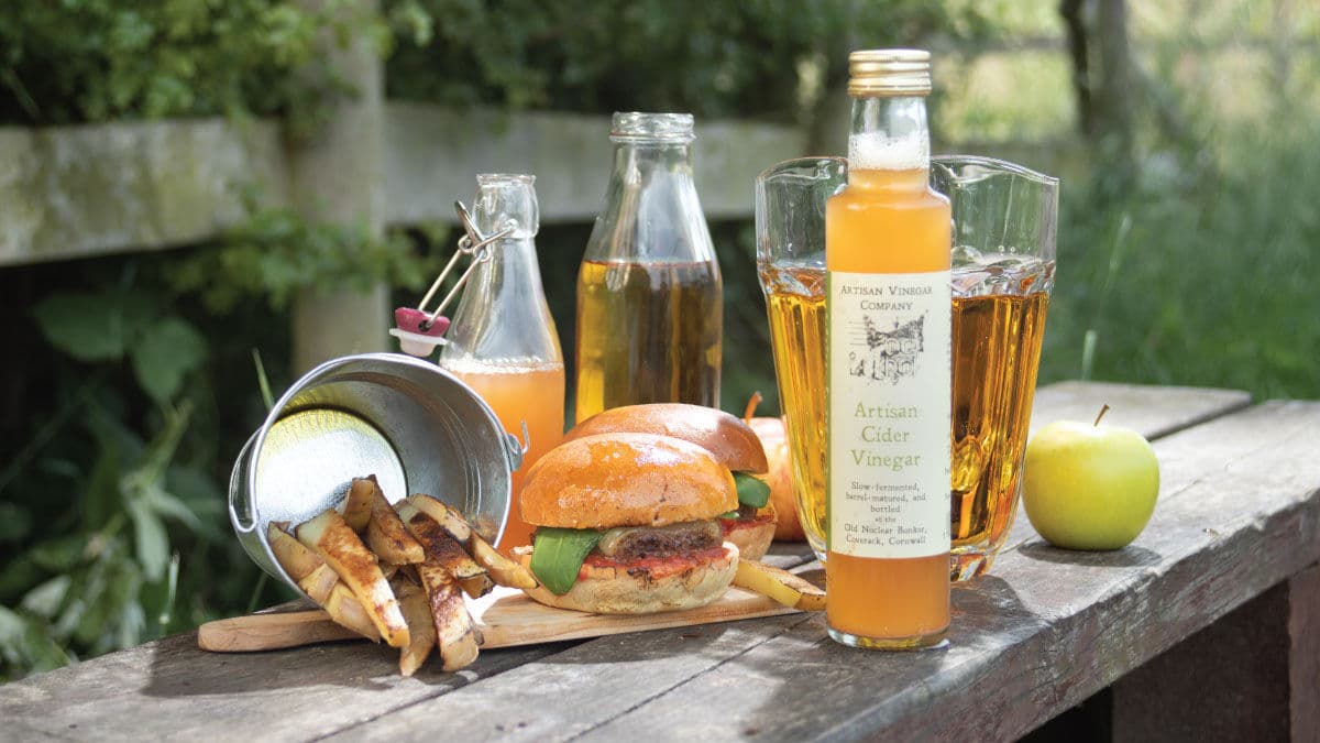 Artisan cider vinegar bottle beside a burger, chips and apple on an outdoor table