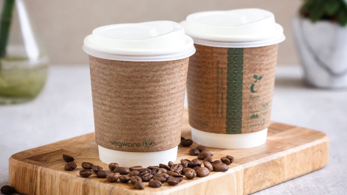Two compostable kraft paper coffee cups with lids on a board with coffee beans